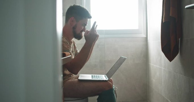 Fun young businessman working on laptop sitting at toilet clenching fist celebrating financial profits doing business in the bathroom.