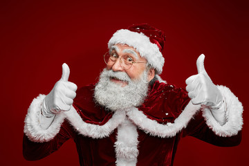 Waist up portrait of classic Santa Claus showing thumbs up while posing against red background in...
