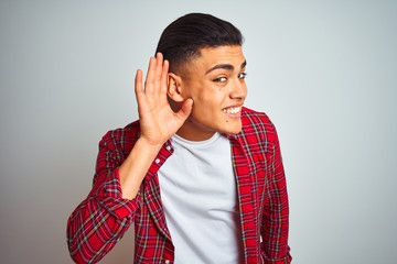 Young brazilian man wearing red shirt standing over isolated white background smiling with hand over ear listening an hearing to rumor or gossip. Deafness concept.