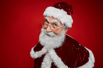 Portrait of smiling Santa Claus looking at camera mischievously while posing against red background in studio, copy space