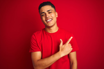 Young brazilian man wearing t-shirt standing over isolated red background cheerful with a smile on face pointing with hand and finger up to the side with happy and natural expression