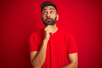 Fototapeta premium Young handsome indian man wearing t-shirt over isolated red background with hand on chin thinking about question, pensive expression. Smiling with thoughtful face. Doubt concept.