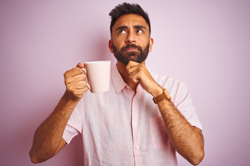 Young indian man wearing shirt drinking cup of coffee standing over isolated pink background serious face thinking about question, very confused idea