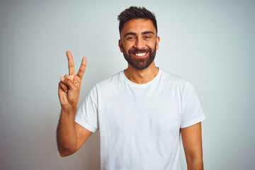 Fototapeta premium Young indian man wearing t-shirt standing over isolated white background showing and pointing up with fingers number two while smiling confident and happy.
