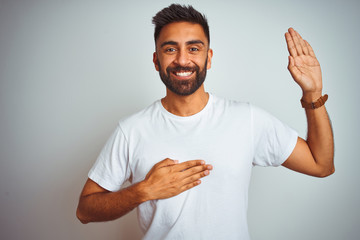 Young indian man wearing t-shirt standing over isolated white background smiling swearing with hand on chest and fingers up, making a loyalty promise oath - obrazy, fototapety, plakaty