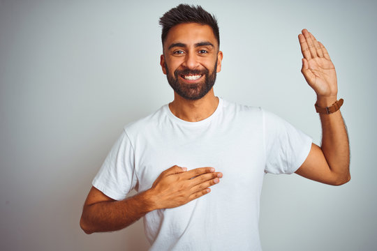 Young indian man wearing t-shirt standing over isolated white background smiling swearing with hand on chest and fingers up, making a loyalty promise oath