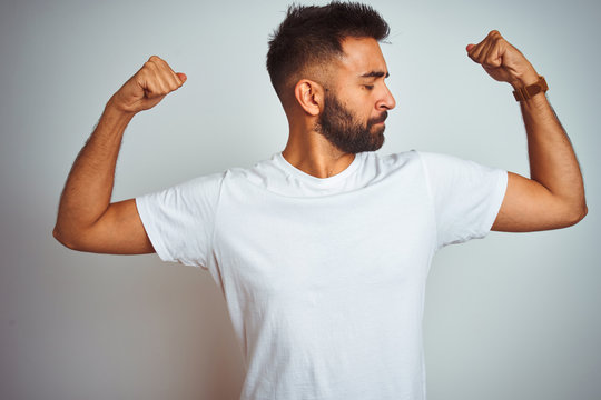 Young indian man wearing t-shirt standing over isolated white background showing arms muscles smiling proud. Fitness concept.