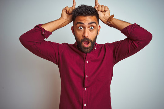 Young indian man wearing red elegant shirt standing over isolated grey background doing funny gesture with finger over head as bull horns