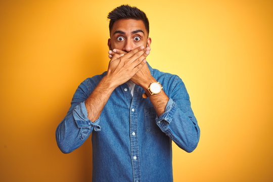 Young indian man wearing denim shirt standing over isolated yellow background shocked covering mouth with hands for mistake. Secret concept.