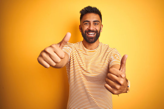 Young indian man wearing t-shirt standing over isolated yellow background approving doing positive gesture with hand, thumbs up smiling and happy for success. Winner gesture.