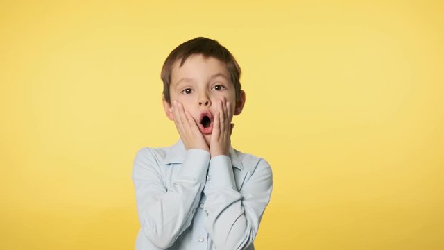 Cute boy kid surprised with hands on his face. Caucasian boy in blue shirt doing silly funny face at yellow background. 4k, UHD 60FPS slow motion