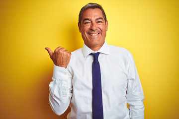 Handsome middle age businessman standing over isolated yellow background smiling with happy face looking and pointing to the side with thumb up.