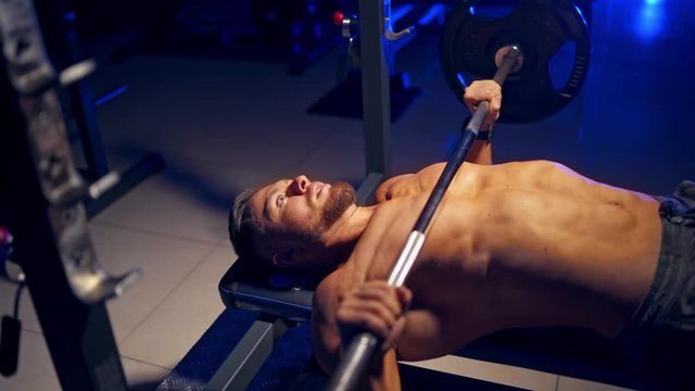 Shirtless young bodybuilder doing bench press for chest. Strong abs. Blue light filter. Video from the side.