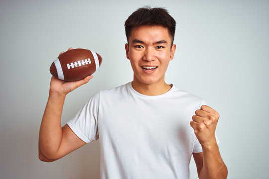 Young asian chinese sportsman holding rugby ball standing over isolated white background screaming proud and celebrating victory and success very excited, cheering emotion