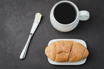 French breakfast. White mug with black coffee and croissant with butter knife on dark stone table. Top view. Coffee break.