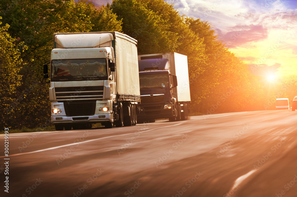 Sticker Trucks with cars on the coutryside road in motion with trees against a sky with a sunset