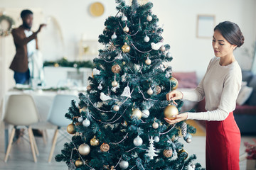 High angle portrait of modern couple preparing for dinner party at home, focus on young woman decorating Christmas tree in foreground, copy space
