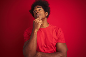 Obraz na płótnie Canvas Young american man with afro hair wearing t-shirt standing over isolated red background with hand on chin thinking about question, pensive expression. Smiling with thoughtful face. Doubt concept.