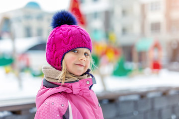 Cute blond kid girl portrait wearing pink knitted hat and sport skiing jacket having fun at playground outdoor during cold winter holidays. Funny playful little child outside. Smiling tricky toddler