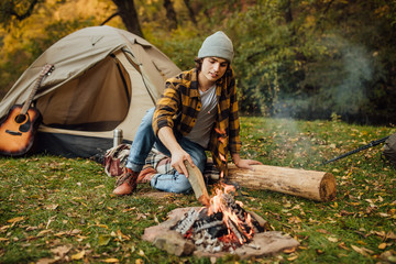 Young attractive man sitting on the log and throws firewood near a tourist tent in the forest.