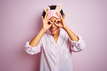Young african american woman wearing pajama and mask over isolated pink background doing ok gesture like binoculars sticking tongue out, eyes looking through fingers. Crazy expression.