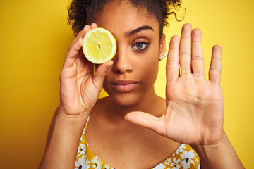 Young african american woman holding slice of lemon over isolated yellow background with open hand doing stop sign with serious and confident expression, defense gesture