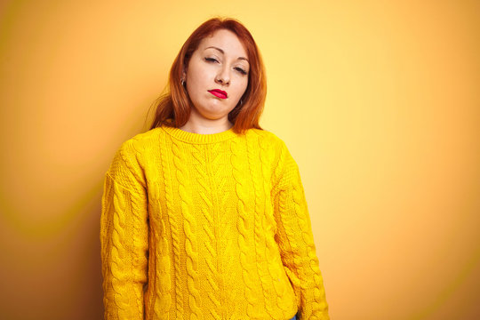 Beautiful redhead woman wearing winter sweater standing over isolated yellow background looking sleepy and tired, exhausted for fatigue and hangover, lazy eyes in the morning.