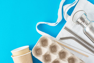 Zero waste concept. Eco friendly reusable items on natural shopping bag. Paper egg tray, coffee cups, aluminum bottle and metal tubes on a blue background