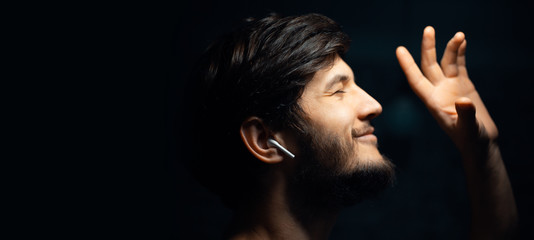Close-up portrait of young hipster man, listen the music with wireless earphones, on black...