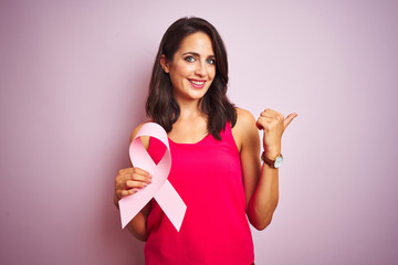 Young beautiful woman holding cancer ribbon over pink isolated background pointing and showing with...