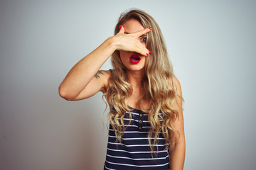 Young beautiful woman wearing stripes t-shirt standing over white isolated background peeking in shock covering face and eyes with hand, looking through fingers with embarrassed expression.