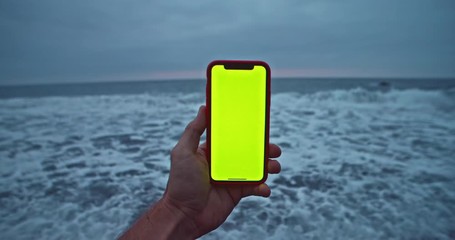 Close-up young man browsing content on mock-up greenscreen smartphone at picturesque seascape. Man using mobile phone on beautiful beach with calm sea in the evening. - Powered by Adobe