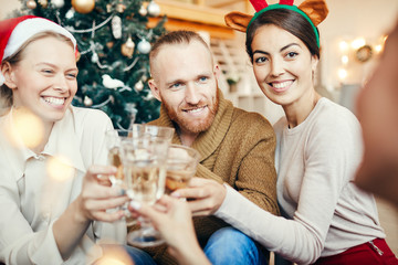 Group of cheerful friends clinking champagne glasses sitting by Christmas tree during party, copy space