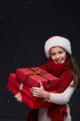 Young beautiful smiling girl in white knitting pullover, red scarf and Santa hat holding gift boxes on a dark background with snowflakes.Winter holidays, Christmas, New Year concept.