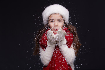 Young beautiful smiling girl in red Santa hat is blowing white snowflakes on a dark background. Xmas fashion model with long straight hair. Winter holidays, Christmas, New Year concept.