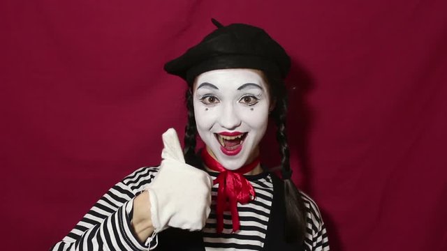 Beautiful girl mime smiles, shows thumb up and nods approvingly while looking at the camera. Beautiful girl in the image of mime shows thumb up while looking at the camera. Portrait on a red backgroun