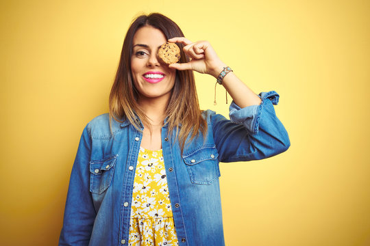 Young beautiful woman eating chocolate chips cookie over yellow background with a happy face standing and smiling with a confident smile showing teeth