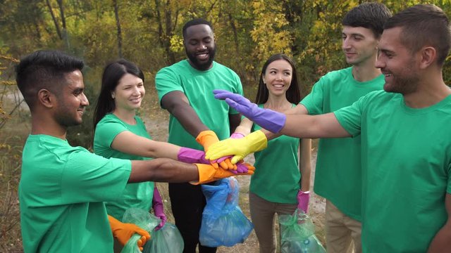 Happy team of diverse activists joining hands in pile and raising arms up while standing together among forest. Joyful multi-ethnic people in green shirts satisfied with garbage collection teamwork