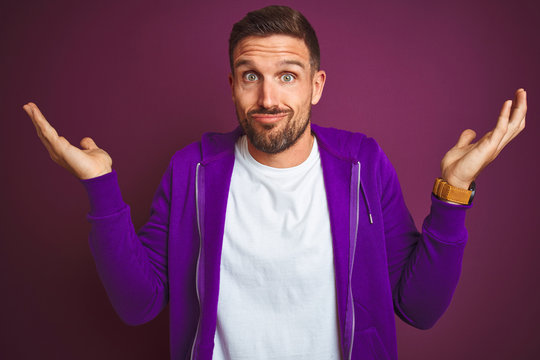 Young fitness man wearing casual sports sweatshirt over purple isolated background clueless and confused expression with arms and hands raised. Doubt concept.