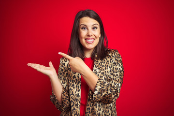 Young beautiful brunette woman wearing a leopard jacket over red isolated background amazed and smiling to the camera while presenting with hand and pointing with finger.