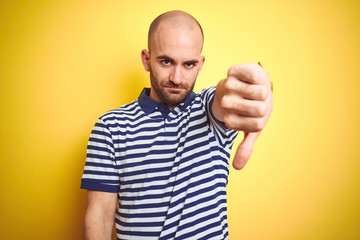Naklejka premium Young bald man with beard wearing casual striped blue t-shirt over yellow isolated background looking unhappy and angry showing rejection and negative with thumbs down gesture. Bad expression.