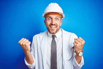 Young handsome engineer man wearing safety helmet over blue isolated background celebrating surprised and amazed for success with arms raised and open eyes. Winner concept.