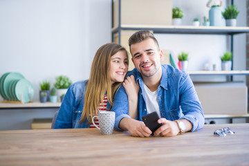 Young couple relaxing drinking a cup of coffee and using smartphone, sitting at new home with cardboard boxes behind them