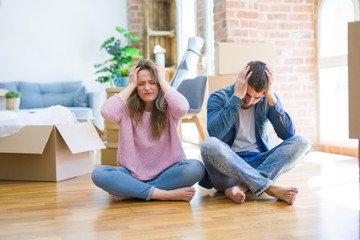 Young beautiful couple moving to a new house sitting on the floor suffering from headache desperate...