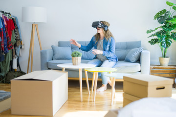 Young blonde woman wearing virtual reality glasses playing a simulation game sitting on the sofa around cardboard boxes moving to a new house