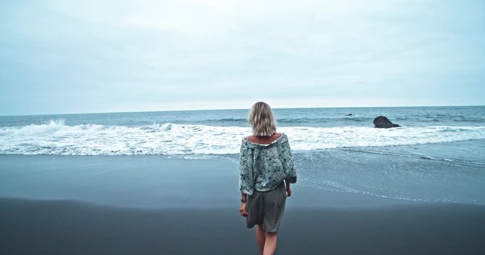 Blonde young insired woman walking on black sand beach into water observing endless wonderful ocean on nice summer day. Freedom and traveling.