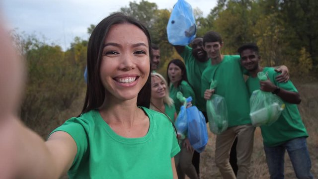 Close-up of smiling asian woman holding cellphone while taking selfie photo of joyful multiracial volunteers. Cheerful group of ecology activists with full garbage bags rejoicing at well-done teamwork