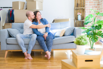 Fototapeta na wymiar Young couple sitting on the sofa arround cardboard boxes moving to a new house looking to side, relax profile pose with natural face with confident smile.
