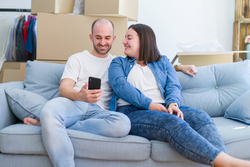 Obraz premium Young couple sitting on the sofa at new home using smartphone, hugging and smiling happy for moving to a new apartment
