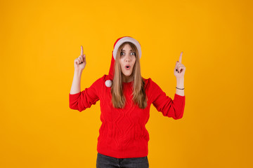 Fototapeta na wymiar excited woman pointing fingers at copy space wearing santa hat and red sweater isolated over yellow
